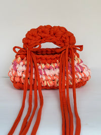 Orange crochet bag with a coral, pink and butter band, a bobbled top handle and long ties tied in bows