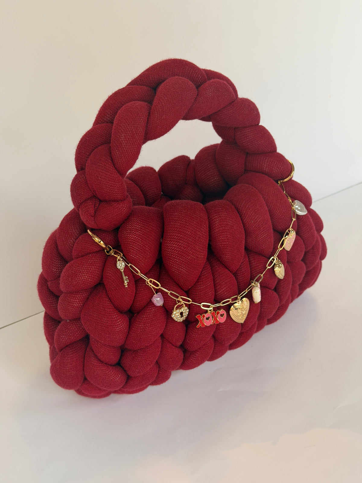 Deep red chunky crochet bag with a braided top handle and a gold charm chain of hearts across the front