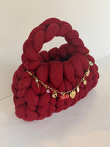 Deep red chunky crochet bag with a braided top handle and a gold charm chain of hearts across the front