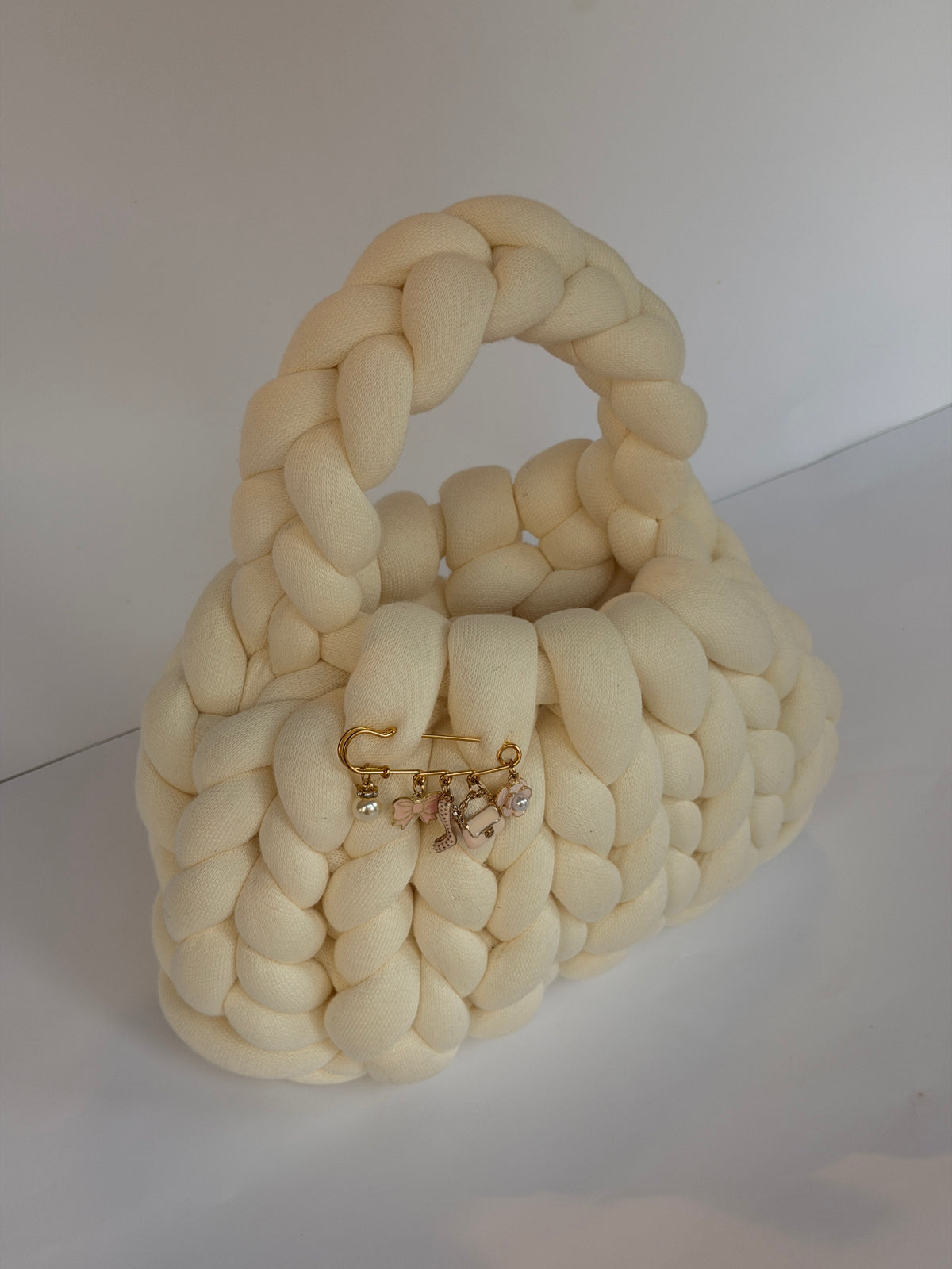 Angled view of a cream crochet bag in jumbo yarn with a gold charm brooch pinned at the front