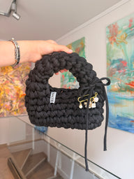 Near-black crochet bag with a rounded top handle, drawstring tie and gold charm pin