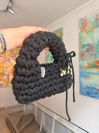 Near-black crochet bag with a rounded top handle, drawstring tie and gold charm pin
