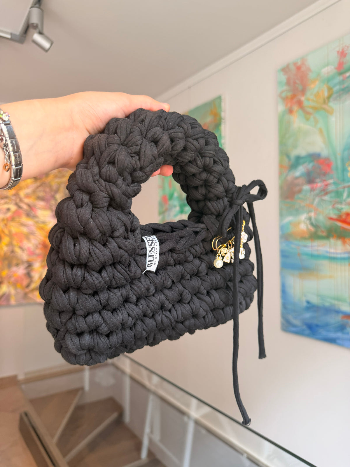 Near-black crochet bag with a rounded top handle, drawstring tie and gold charm pin
