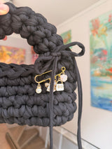 Near-black crochet bag with a rounded top handle, drawstring tie and gold charm pin