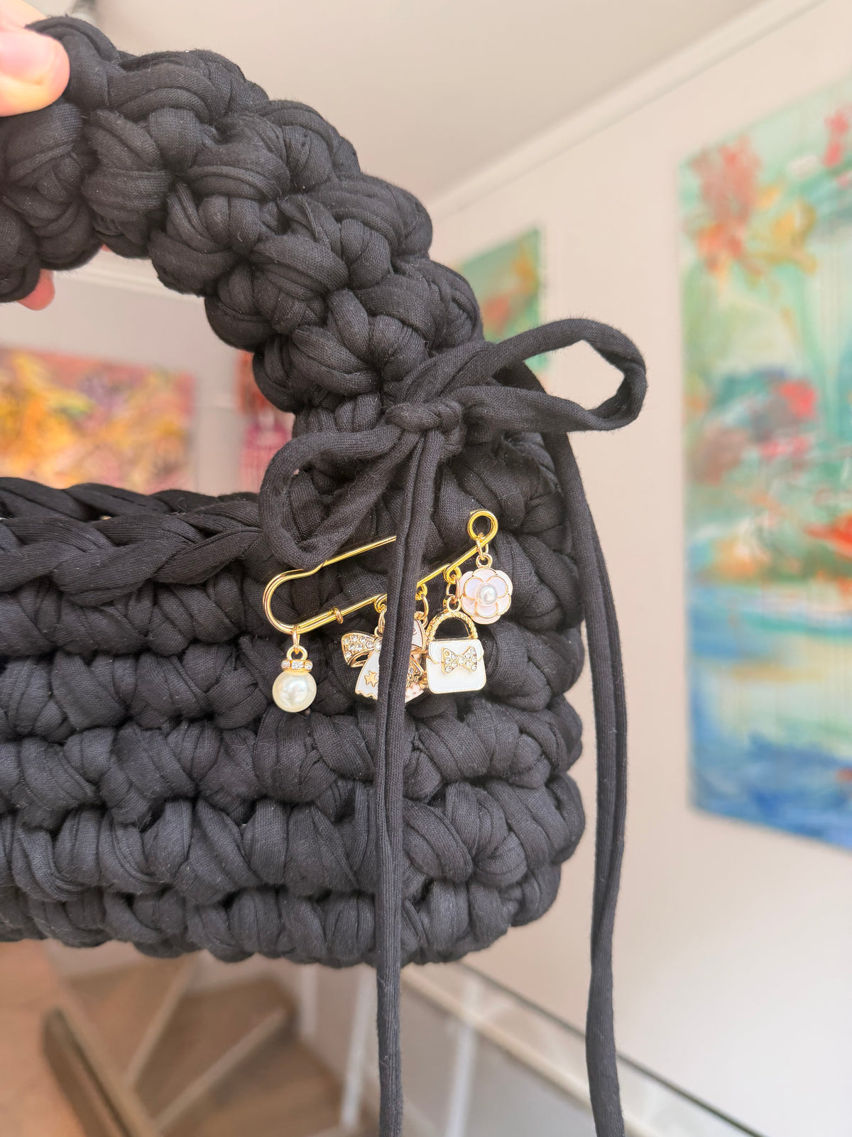 Near-black crochet bag with a rounded top handle, drawstring tie and gold charm pin